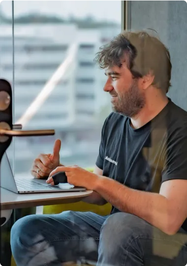 Un homme assis dans une salle vitrée, travaillant sur un ordinateur portable, portant un t-shirt noir avec un design discret, concentré sur sa tâche dans un environnement moderne.