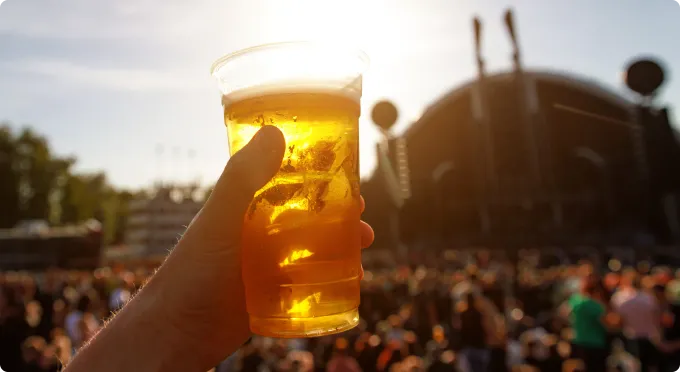 Main tenant un verre de bière dans un festival en plein air, avec une foule et une scène en arrière-plan.