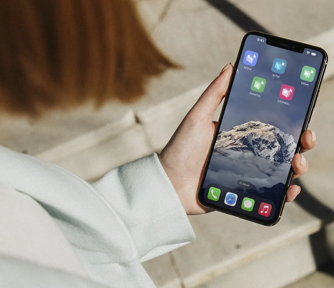 Photo of a hand holding a smartphone displaying a home screen with kSuite application icons, against a background image of snow-capped mountains.