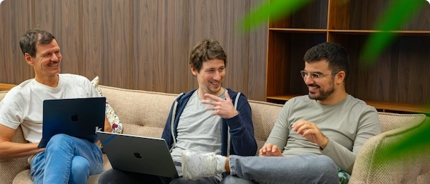 Tres compañeros sonrientes sentados en un sofá con portátiles, en un ambiente de oficina relajado.