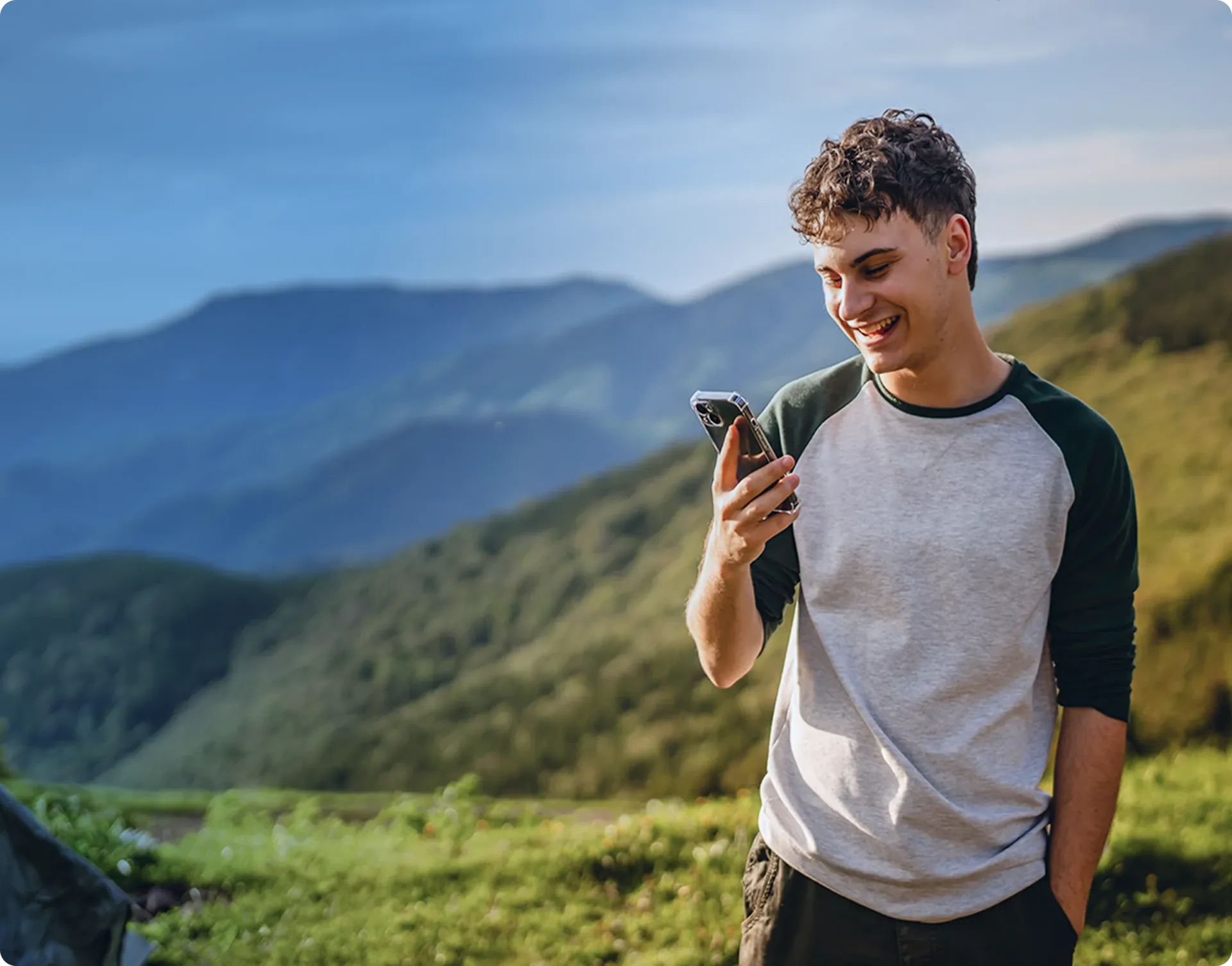 A young man carrying a backpack, dressed in a green jumper, using his smartphone while standing in a natural landscape with hills and trees in the background.