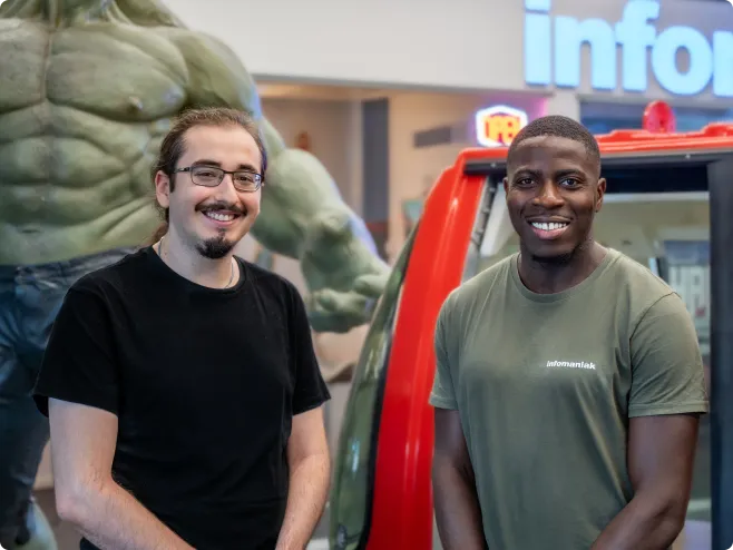 Deux membres de l'équipe de support souriants, l'un portant un t-shirt Infomaniak, posant devant une statue imposante et un télécabine rouge, représentant un service d'assistance local et accessible.