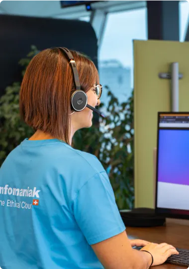 Une femme portant un casque avec micro et un t-shirt bleu Infomaniak, travaillant devant un écran d'ordinateur dans un environnement lumineux avec des plantes en arrière-plan.