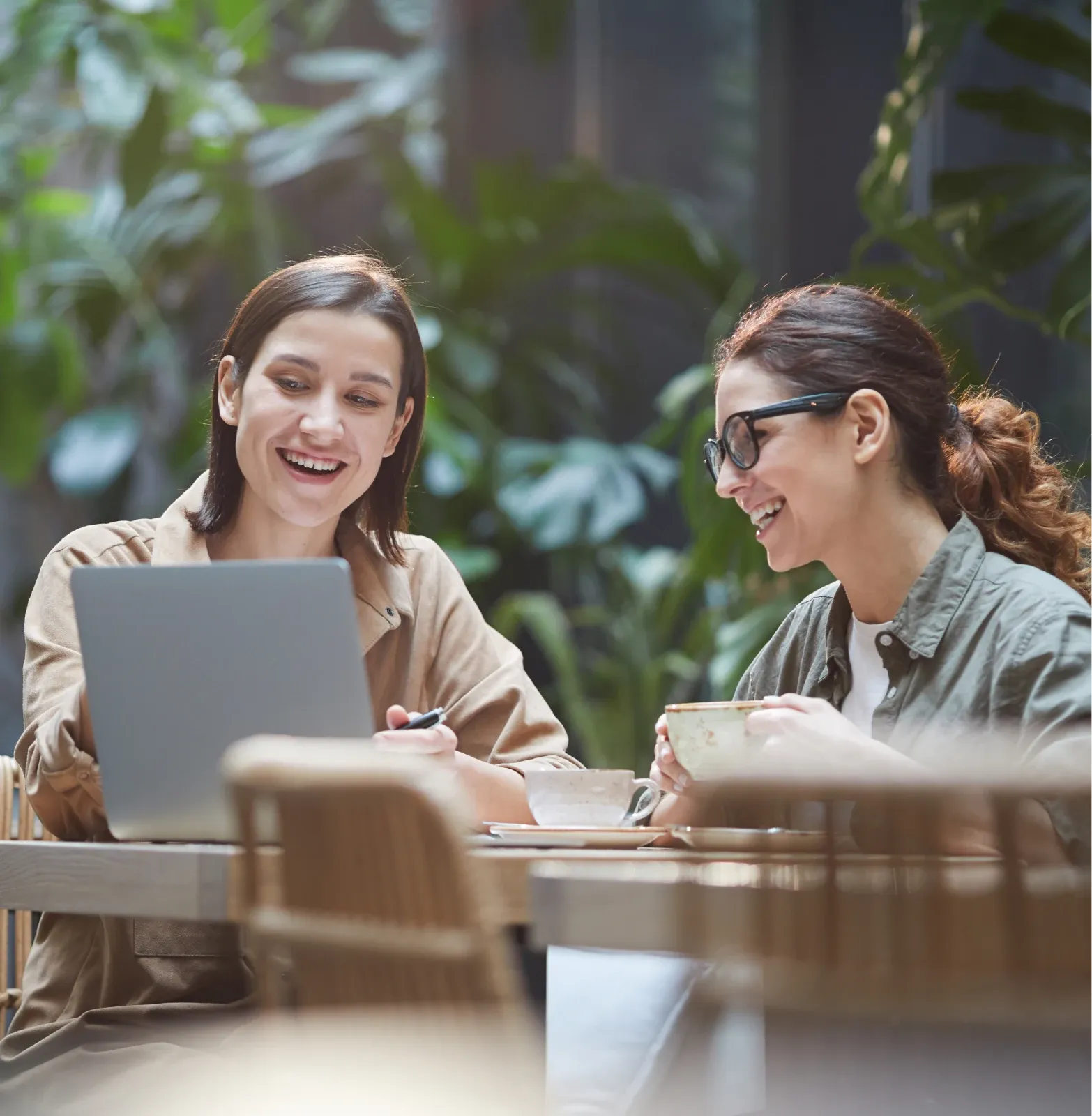 Two smiling women working together on a laptop, seated at a table in a bright, green space surrounded by plants.