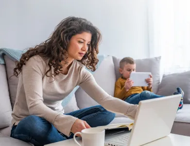 A woman working on a laptop in a living room, with a child playing on a tablet in the background, illustrating a secure, privacy-friendly environment.